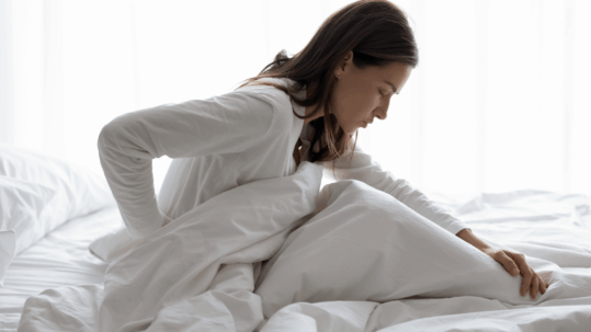 A woman sits up in bed, holding her lower back in discomfort, illustrating the kind of chronic back pain that can wake you at night or limit daily tasks, signaling the need for specialist care.