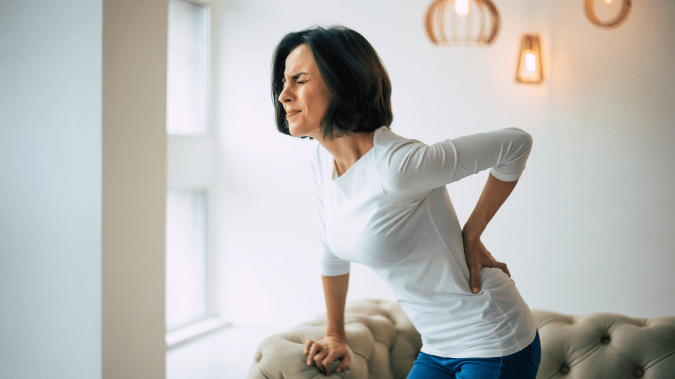A woman with a pained expression, clutching her lower back while leaning on a couch, illustrating the common symptoms of spinal stenosis such as pain and discomfort with standing or walking.