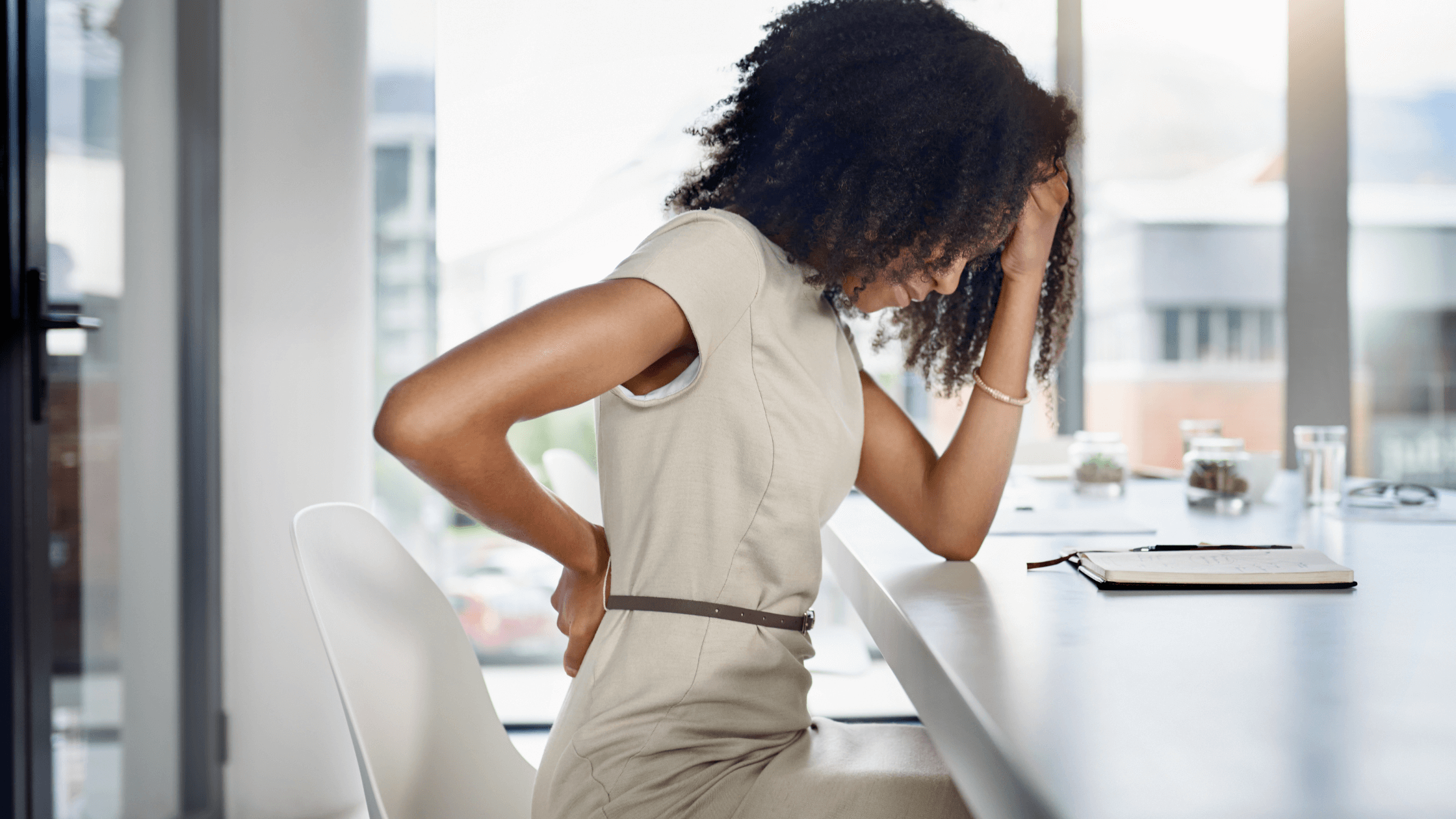 A woman sitting at an office desk, clutching her lower back in discomfort, illustrating pain that worsens while sitting, often linked to conditions like disc herniation