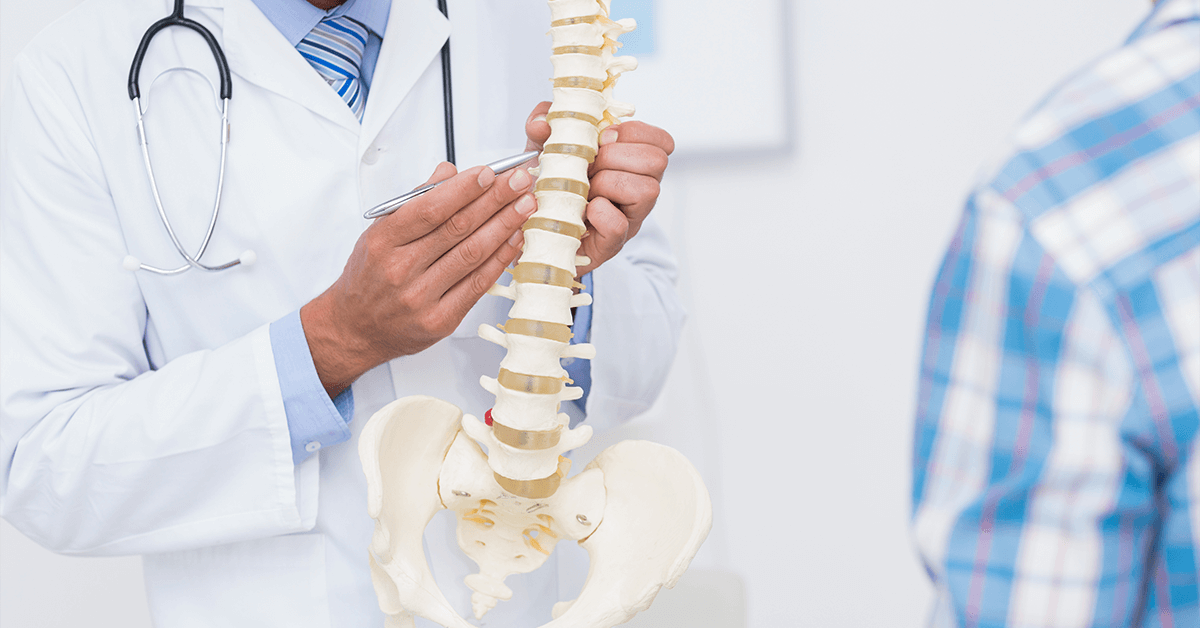 A doctor in a white coat uses a pen to point out a feature on an anatomical model of the lumbar spine and pelvis to a patient, illustrating a pain management consultation and patient education about their spinal condition.