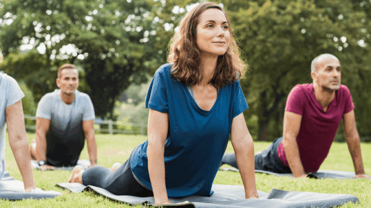 A group of adults practices gentle yoga outdoors on mats, performing a cobra pose with a calm expression, symbolizing physical therapy and gentle movement for pain management and improved flexibility.